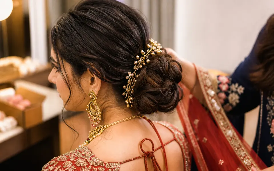 Side and back view of an Indian bride with fine hair styled into a softly textured low bun, while a stylist secures pins and a dupatta.