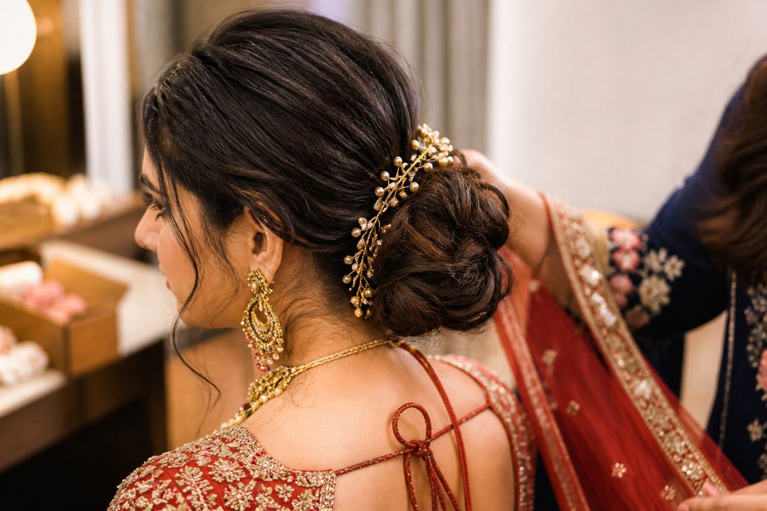 AI-generated illustration: Side and back view of an Indian bride with fine hair styled into a softly textured low bun, while a stylist secures pins and a dupatta.