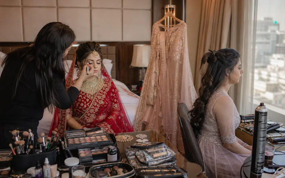 Indian bride and her sister getting ready in a Delhi hotel room, with a makeup artist working on the bride while the sister waits with her hairstyle partly done.