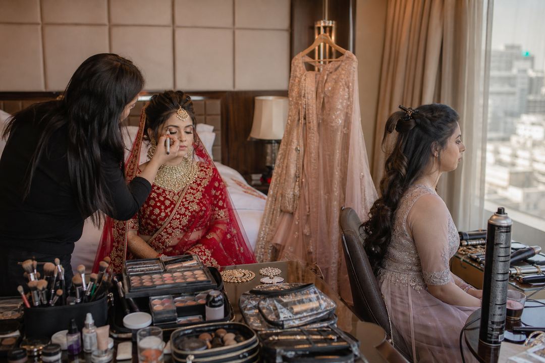 AI-generated illustration: Indian bride and her sister getting ready in a Delhi hotel room, with a makeup artist working on the bride while the sister waits with her hairstyle partly done.